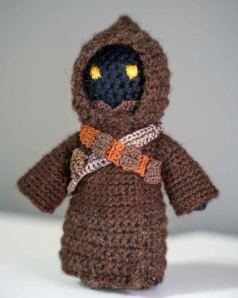 Peut inclure: Une figurine &agrave; capuche en crochet marron avec des yeux jaunes et une ceinture en crochet marron et orange.