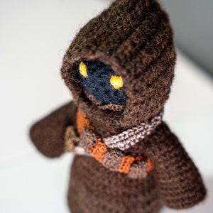 Peut inclure: Un jouet en peluche crochet&eacute; marron avec une robe &agrave; capuche et des yeux jaunes. Le jouet est fait de fil doux et a un visage d&eacute;taill&eacute;.