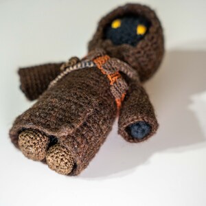Peut inclure: Un jouet en peluche crochet&eacute; marron avec une robe &agrave; capuche et des yeux noirs. Le jouet est allong&eacute; sur le dos, les bras tendus.
