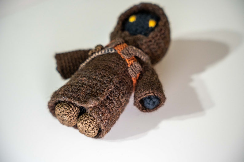 Peut inclure: Un jouet en peluche crochet&eacute; marron avec une robe &agrave; capuche et des yeux noirs. Le jouet est allong&eacute; sur le dos, les bras tendus.