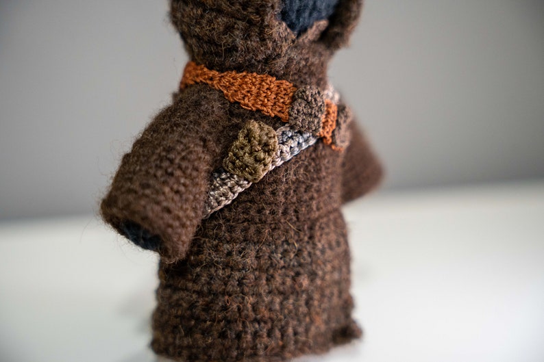 Peut inclure: Un jouet en peluche crochet&eacute; marron avec une ceinture crochet&eacute; marron, orange et verte. Le jouet porte une robe &agrave; capuche marron.