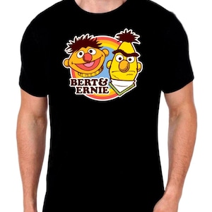 May include: A black short-sleeved t-shirt featuring a graphic print of the characters Bert and Ernie. The design includes a colourful rainbow circle background, the text BERT & ERNIE in brown, and the two characters in their signature colours.