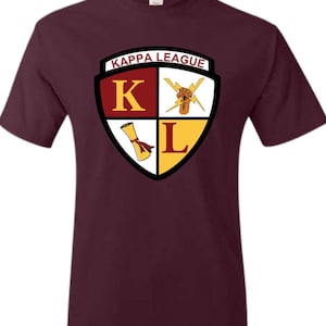 Può includere: T-shirt bordeaux con un design a scudo. Lo scudo presenta le parole "KAPPA LEAGUE" in alto e le lettere "K" e "L" in rosso e oro. Lo scudo include anche un fulmine e una pergamena.