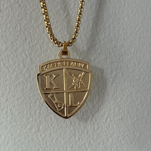 Puede incluir: Un collar dorado con un colgante en forma de escudo. El colgante presenta las letras "K" y "L" en cuadrantes separados, junto con otros símbolos. La cadena es de estilo eslabón cuadrado. El colgante tiene las palabras "KAPPA LAMBDA" en la parte superior.