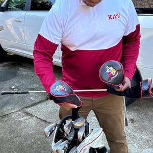 May include: A person wearing a white and burgundy golf shirt with the letters "KAY" embroidered on the chest. The person is holding two black golf club head covers with red trim and a crest featuring a shield and a crown. The head covers are both embroidered with the letters "KAY".