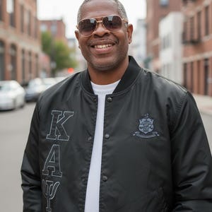 Puede incluir: Una chaqueta bomber negra con las letras "K A Ψ" y un escudo. La chaqueta se lleva sobre una camiseta blanca. La persona lleva gafas de sol y sonríe.