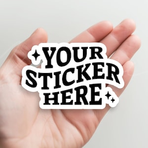 May include: A white sticker with black text that reads "YOUR STICKER HERE" with two small stars on either side of the text. The sticker is being held in a person's hand.