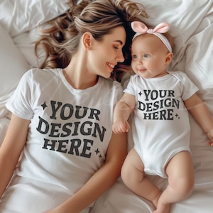 May include: A white t-shirt with the text "YOUR DESIGN HERE" in black lettering. A baby is wearing a white onesie with the same text. The baby is wearing a pink headband. The mother and baby are lying on a white bed.