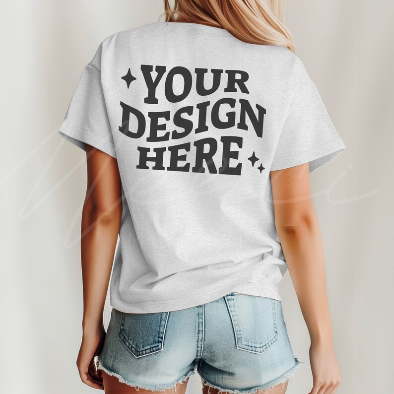 K&ouml;nnte beinhalten: Ein wei&szlig;es T-Shirt mit dem Text "YOUR DESIGN HERE" in schwarzer Schrift auf der R&uuml;ckseite. Das Shirt wird von einer Person mit langen blonden Haaren getragen und zu Jeans-Shorts kombiniert.