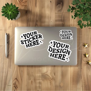 Pode incluir: Três adesivos preto e branco em um laptop prateado. Os adesivos dizem "Your Sticker Here", "Your Design Here" e "Your Design Here".