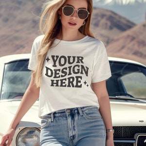 May include: A white t-shirt with the text "YOUR DESIGN HERE" in black lettering. The shirt is being worn by a person with long blonde hair and sunglasses. The person is standing in front of a vintage car.