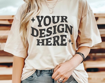 May include: A beige t-shirt with the text "YOUR DESIGN HERE" printed in black. The shirt is being worn by a person with long blonde hair.