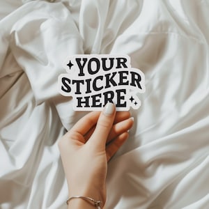 May include: A white sticker with black text that reads "YOUR STICKER HERE" with two small stars on either side of the text. The sticker is being held by a hand.