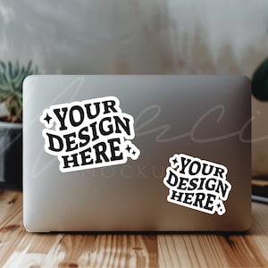 May include: A silver laptop computer with two white stickers on the lid. The stickers have black text that reads "Your Design Here" with a star on each side.