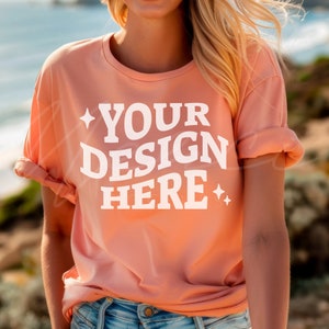 May include: A peach colored t-shirt with the text "YOUR DESIGN HERE" in white. The shirt is being worn by a person with long blonde hair.