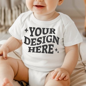 以下が含まれることがあります： 白い半袖のベビーロンパースで、黒い文字で「YOUR DESIGN HERE」と書いてあり、文字の両側に小さな星が2つあります。