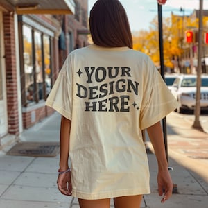 Puede incluir: Camiseta amarillo claro con el texto "YOUR DESIGN HERE" en gris oscuro, con estrellas. La camiseta es de corte holgado, ideal para diseños personalizados. La imagen está tomada al aire libre.