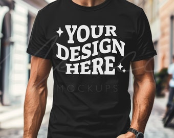 Puede incluir: Una camiseta negra con el texto "YOUR DESIGN HERE" en letras blancas. La camiseta la lleva puesta una persona que está de pie en un entorno urbano.