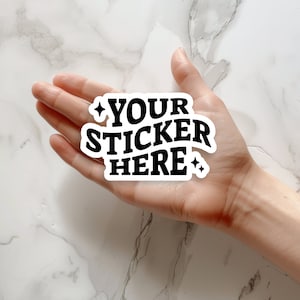 Può includere: Un adesivo bianco con testo nero che recita "YOUR STICKER HERE" con due piccole stelle su entrambi i lati del testo. L'adesivo è tenuto nella mano di una persona.