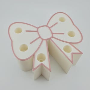 May include: A white bow-shaped desk organizer featuring five circular holes for holding pens or pencils. The bow design is outlined with a thin, raised pink border that follows the shape of the loops and the two downward-hanging ribbon tails.