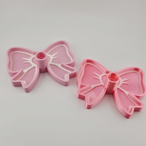 May include: Two pink bow-shaped candle holders. Each holder has a central cylinder for a candle and a detailed bow design. The bows are a solid pink with white accents, set against a white background.