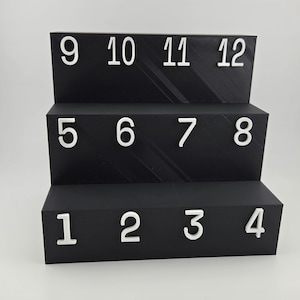 May include: A black three-tiered display stand with a textured surface. Each tier features white raised numbers. The bottom tier displays 1, 2, 3, 4. The middle tier displays 5, 6, 7, 8. The top tier displays 9, 10, 11, 12 in a clean, sans-serif font.