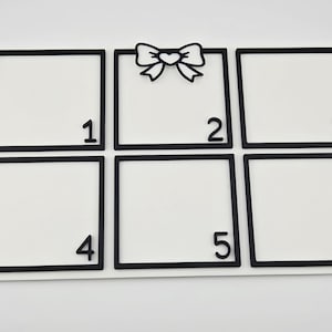 May include: A white display board with six black-framed squares. A black bow decoration sits above the second square. Each square is numbered 1 to 6 in black. The board is designed for displaying photos or small items, possibly for a craft project.