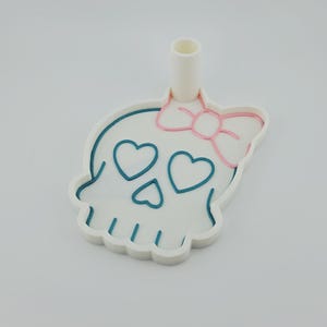 May include: A white plastic skull-shaped cookie cutter featuring a pink bow detail on the top right and teal heart-shaped eye sockets and nose. The cutter includes a vertical cylindrical handle attached to the top for ease of use.