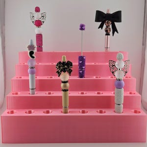 May include: Six decorative pens with various bead and charm toppers, including bows, wings, and floral shapes, displayed in a tiered pink plastic stand. The pens feature diverse colors like pink, purple, black, and tan with textured and smooth beads.