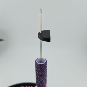 May include: A metal needle tool featuring a textured purple handle with a diamond-patterned grip, a light pink cylindrical base, and a small black semi-circular component attached to the upper shaft, positioned upright on a black circular stand.