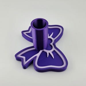 May include: A purple 3D printed stand featuring a bow-shaped base with a light purple outline and a central cylindrical holder. The object has a smooth finish and a distinct layered texture characteristic of additive manufacturing processes.