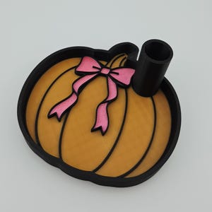May include: A pumpkin-shaped tray with a black outer border and a textured orange interior. A pink bow is attached to the centre of the pumpkin. A black cylindrical holder is positioned on the right side of the tray, extending upwards from the base.