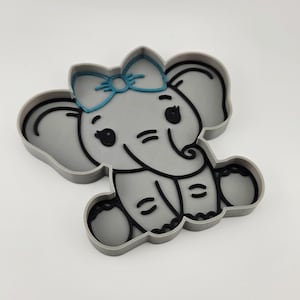 Op de afbeelding: Een grijze koekjesvorm in de vorm van een olifant met een blauwe strik op het hoofd. Het ontwerp heeft zwarte omlijningen voor de ogen, oren, slurf en voeten, met een verhoogd grijs oppervlak dat het lichaam vormt. De vorm heeft een dik, stevig frame.