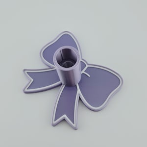 May include: A lavender-colored bow-shaped object featuring a central cylindrical holder. The bow has a raised white outline along its edges, and the cylindrical component is positioned vertically in the center of the bow structure.