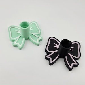 May include: Two bow-shaped candle holders. One is light green with white trim, and the other is black with pink trim. Each has a cylindrical holder in the center for a candle. The candle holders are on a white background.