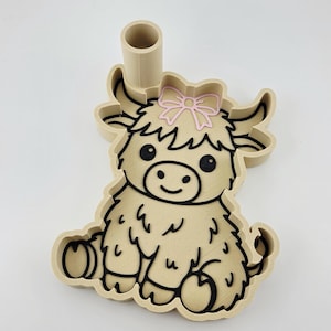 Puede incluir: Un cortador de galletas beige impreso en 3D con forma de vaca Highland con un lazo rosa en la cabeza. El cortador presenta contornos negros para los rasgos faciales, el pelaje y las pezuñas, e incluye un mango cilíndrico en el borde superior.