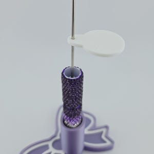May include: A vertical metal needle tool with a textured purple handle, featuring a white circular tag attached to the shaft. The tool is inserted into a light purple base shaped like a bow with a white outline, providing a stable upright stand.