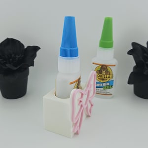 May include: A white rectangular glue bottle holder featuring a pink bow accent on the front. One bottle of Gorilla Super Glue Gel with a blue cap is inserted into the holder, while a second bottle with a green cap stands behind it.