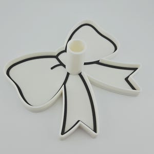May include: A white candle holder shaped like a bow with a central cylindrical opening. The bow features a distinct black outline along its edges and center, providing a high-contrast graphic appearance against the solid white surface.