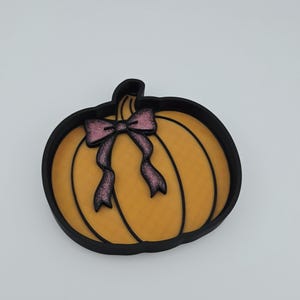 May include: A pumpkin-shaped tray with a black outer rim and a textured orange interior. The pumpkin features black vertical lines and a decorative pink glitter bow attached to the stem area. The tray has a flat base and raised edges.