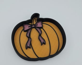 Puede incluir: Una bandeja con forma de calabaza con un borde exterior negro y un interior naranja texturizado. La calabaza presenta líneas verticales negras y un lazo decorativo de purpurina rosa unido a la zona del tallo. La bandeja tiene base plana.