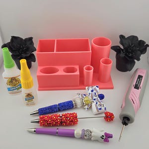 May include: A coral desk organizer, two Gorilla Super Glue bottles, a pink and gray cordless rotary tool, and three decorative pens. One pen has blue rhinestones, one has red flowers, and one has a white ghost figure with a purple bow.