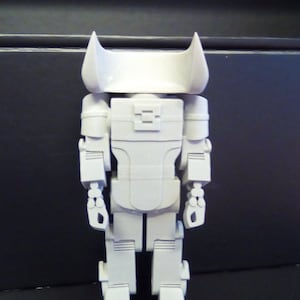 Könnte beinhalten: Ein hellgraues Roboter-Spielzeug mit einem blockartigen Design. Der Roboter hat einen rechteckigen Körper, Arme und Beine, mit einem Kopf mit zwei spitzen Ohren. Das Spielzeug besteht aus einem festen, glatten Material.