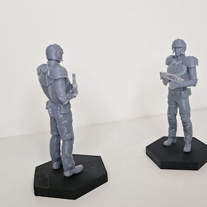 May include: Two grey 3D printed figurines of futuristic soldiers wearing helmets and armour. One soldier is holding a weapon, while the other is holding a device. Both figurines are standing on hexagonal bases.
