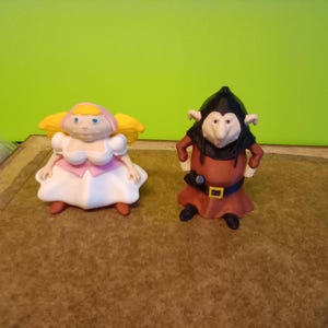 May include: Two plastic figurines standing side by side. The left figure has yellow hair, a pink bodice, and a white skirt. The right figure wears a black hood, a brown tunic with a black belt, and a yellow buckle, featuring large ears and a pointed nose.