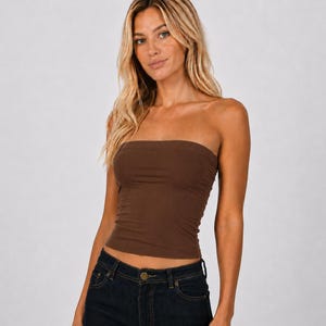 May include: A solid dark brown strapless tube top made of a smooth, stretchy fabric. The garment features a straight-across neckline and a fitted silhouette that ends at the waist, paired with dark blue denim jeans with visible metal buttons.