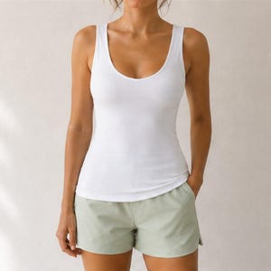 May include: A solid white sleeveless tank top with a scoop neckline paired with light sage green shorts. The tank top features a fitted silhouette and a simple hemline, while the shorts have a relaxed fit and a clean, unadorned appearance.