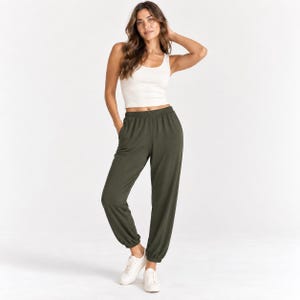 Könnte beinhalten: Ein cremefarbenes, geripptes Tanktop kombiniert mit olivgrüner Jogginghose mit elastischem Bund und Bündchen an den Knöcheln, getragen mit weißen Turnschuhen. Das Outfit besteht aus einem ärmellosen Oberteil mit Rundhalsausschnitt und einer lockeren Hose.