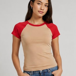 Peut inclure: Un t-shirt à manches raglan courtes présentant un torse de couleur beige uni avec des manches et un col rouge vif. Le vêtement est doté d'un col rond et d'une coupe ajustée, associé à un jean en denim bleu classique avec des rivets métalliques.
