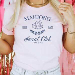 May include: A pink t-shirt with a blue graphic design that says "Mahjong Social Club Tiles & Tea" with a drawing of mahjong tiles and the words "Dot" and "Bam" above and below the tiles.